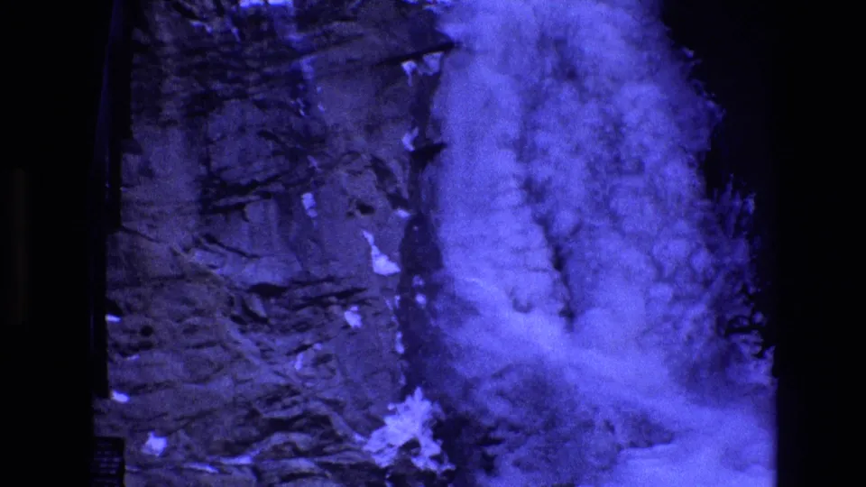 1973 Colorado 8mm frame showing a blue-tinted waterfall cascading over a rough mountain rock face with patches of snow or ice and mist at the base.