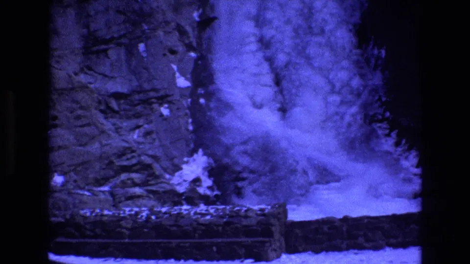 1973 Colorado 8mm frame showing a close view of blue-tinted water and spray over a rugged cliff face, with bright snow or ice pooled along the base.