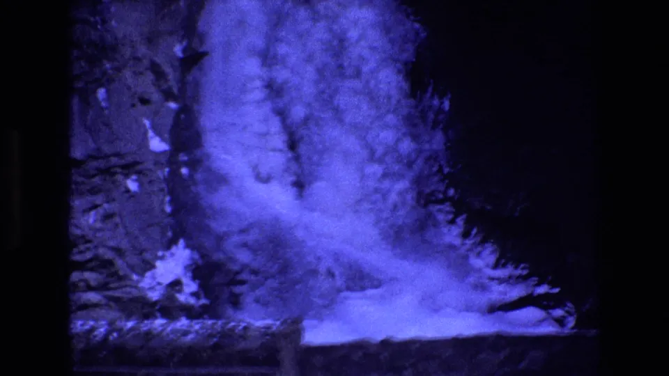 1973 Colorado 8mm frame of a blue-tinted mountain waterfall dropping along a shadowed rock wall, with snow or ice at the bottom and visible film-frame edges.