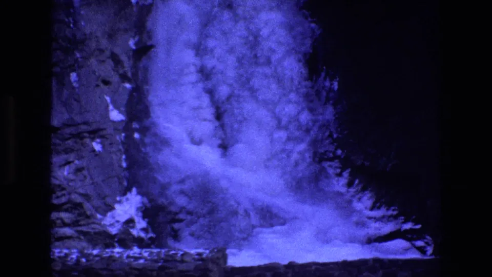 1973 Colorado home-movie frame of the frozen waterfall base, with bright blue ice and snow against a dark rugged rock wall.