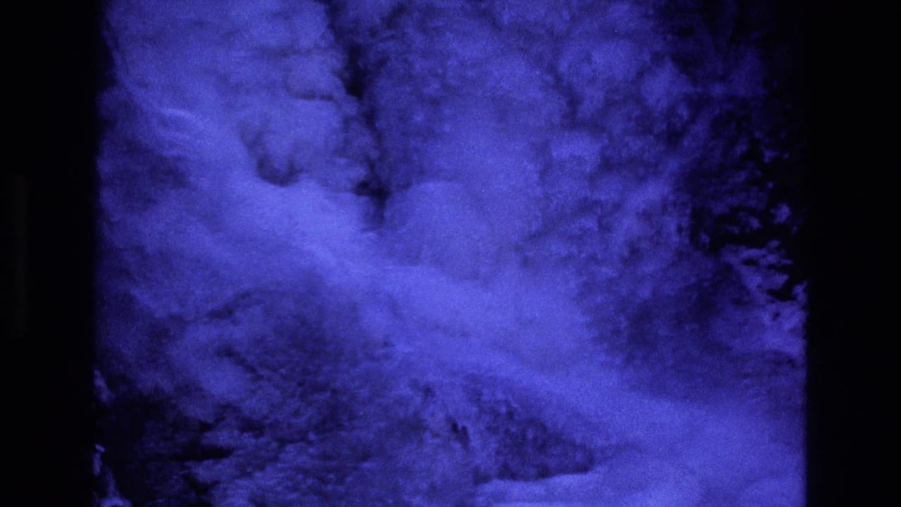 1973 Colorado 8mm frame showing blue-filtered snow or powder moving across a dark rocky mountain slope with heavy grain and projected-film borders.