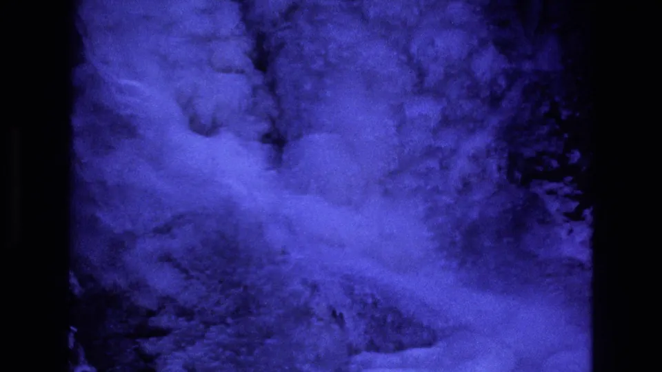 1973 Colorado 8mm frame showing blue-filtered snow or powder moving across a dark rocky mountain slope with heavy grain and projected-film borders.