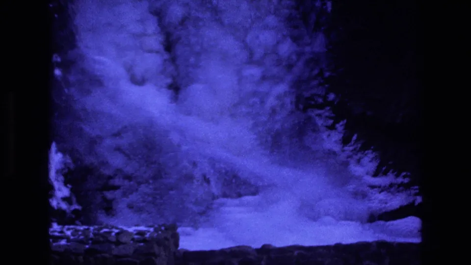 1973 Colorado 8mm frame showing a brighter blue snow drift or flow near the base of a dark slope, with dense grain and strong black borders from the film scan.