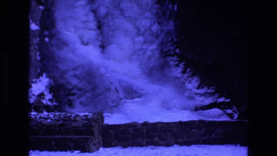 Blue-cast home-movie frame from Colorado showing ice and moving water below a frozen waterfall, with trees and a low ledge in the foreground.