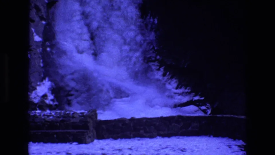 1973 Colorado 8mm frame showing a frozen waterfall in a snowy wooded area, with a dark tree line and rough stone or log-edged ledge.