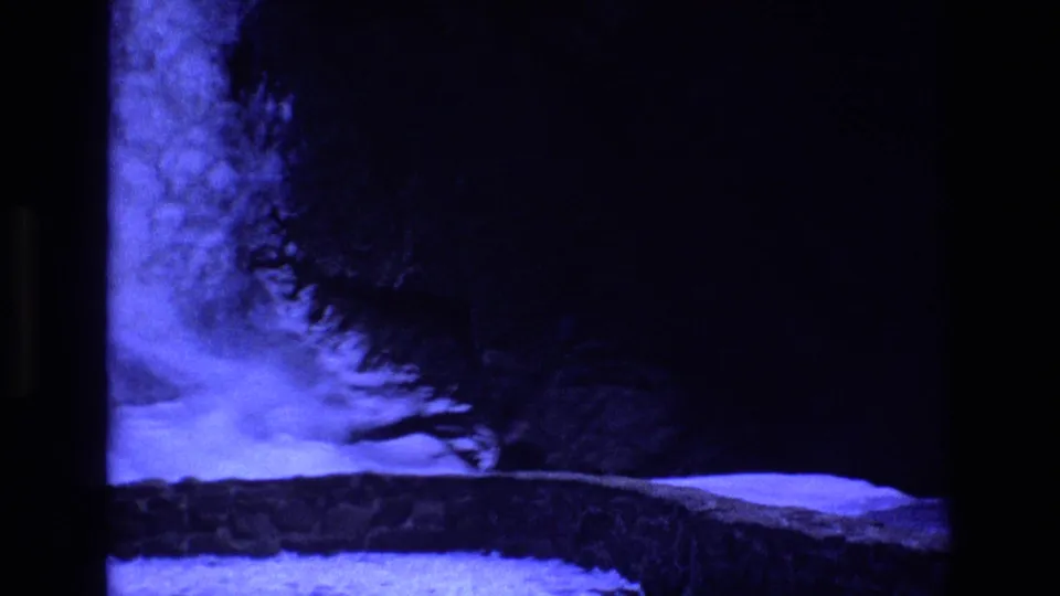Colorado 1973 8mm frame showing a blue-lit rocky ledge or cave-like wall with deep shadows and black frame edges, before the model rocket is clearly centered.