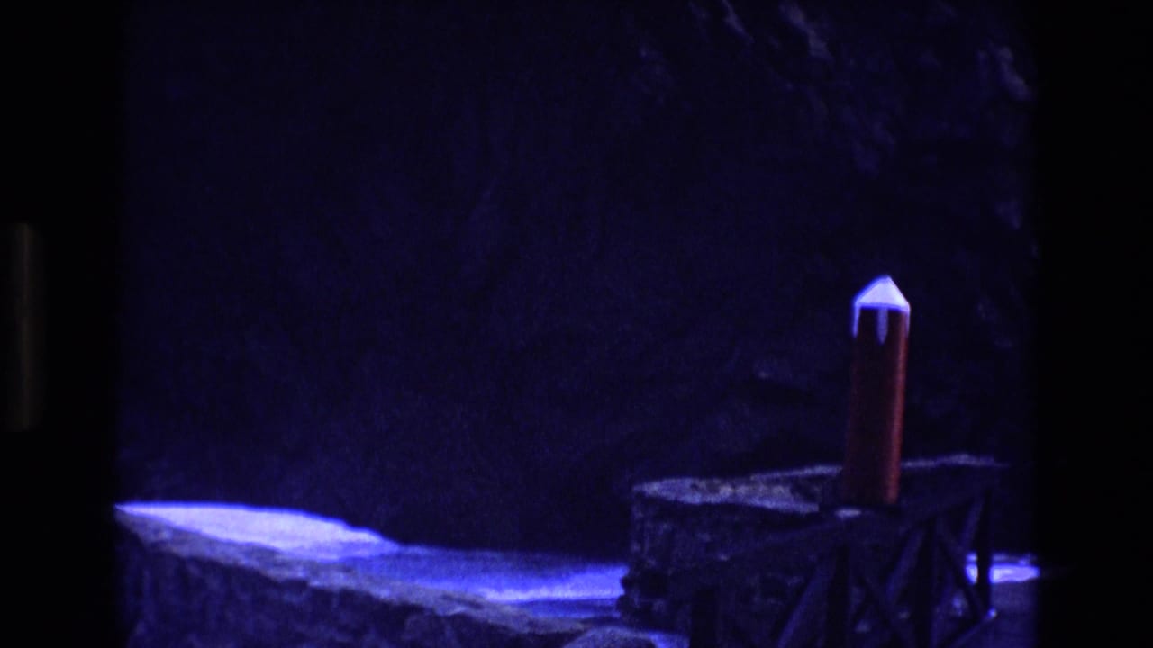 Red model rocket with white nose cone standing on a small wooden structure in blue light.
