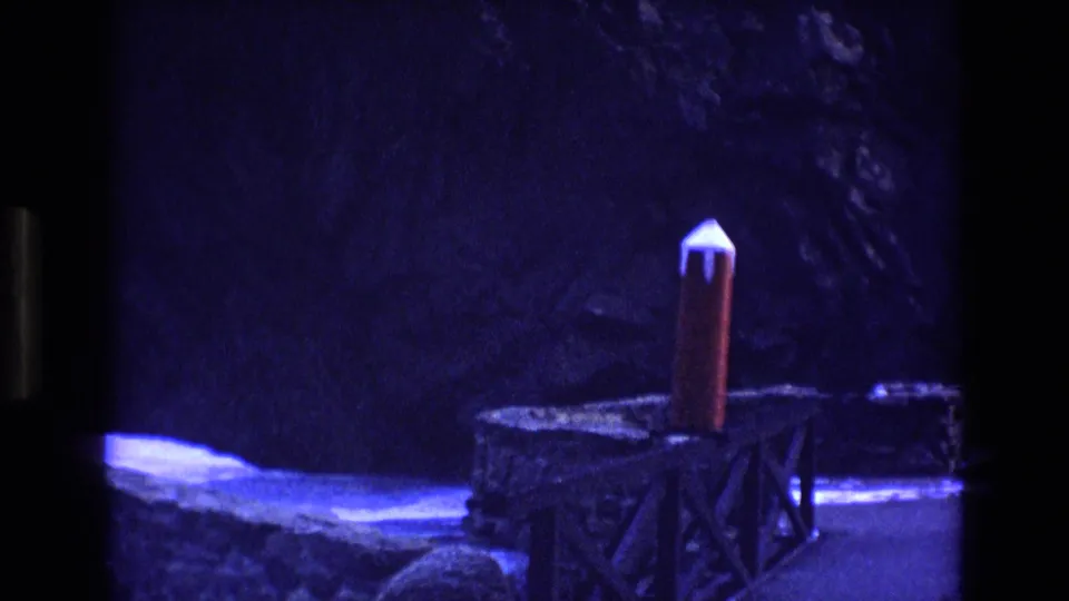 Colorado 1973 8mm frame showing the red-and-white model rocket more centrally on a wooden platform, with rough stone ledges and heavy blue shadow around it.