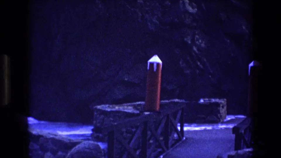 Colorado 1973 8mm frame showing the red model rocket standing on a wooden support near rough stone ledges, with blue illumination and black frame edges visible.