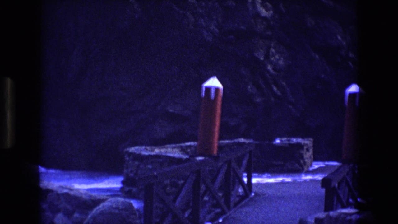 1973 Colorado 8mm frame showing a rocky visitor walkway with wooden railings, red-topped posts, and dark cliffside surroundings under a strong blue-purple cast.