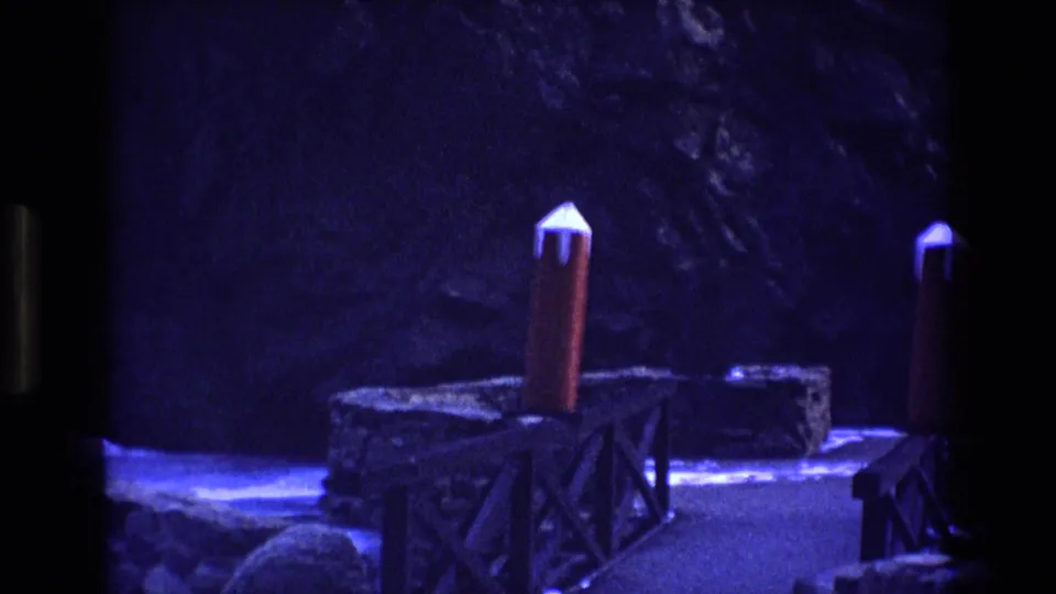 1973 Colorado 8mm frame showing a rocky visitor walkway with wooden railings, red-topped posts, and dark cliffside surroundings under a strong blue-purple cast.
