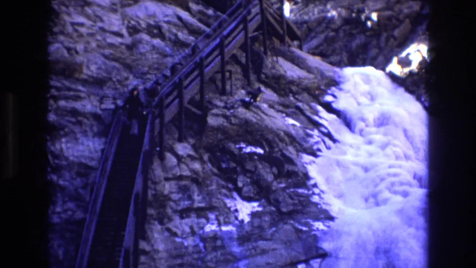 1973 Colorado 8mm frame showing a steep wooden-railed stairway cut into rock beside a bright rushing waterfall or cascade.