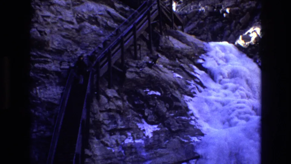 1973 Colorado 8mm frame showing a distant person in dark clothing walking on steep railed stairs beside rushing water and rugged rock.