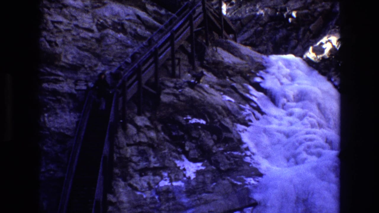 1973 Colorado 8mm frame showing a dark wooden-railed mountain trail beside a rocky cliff, with white water cascading over snow-dusted stone.