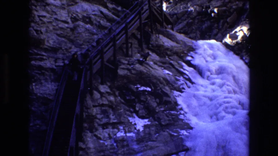 1973 Colorado 8mm frame showing a dark wooden-railed mountain trail beside a rocky cliff, with white water cascading over snow-dusted stone.