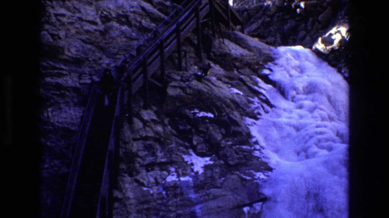 Waterfall pours over dark rock near a wooden railing on a mountain path.