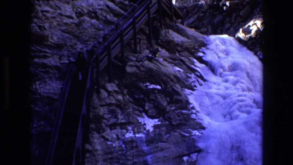 1973 Colorado home-movie frame with a strong waterfall spilling down a rocky wall beside a shadowed wooden-railed walkway.