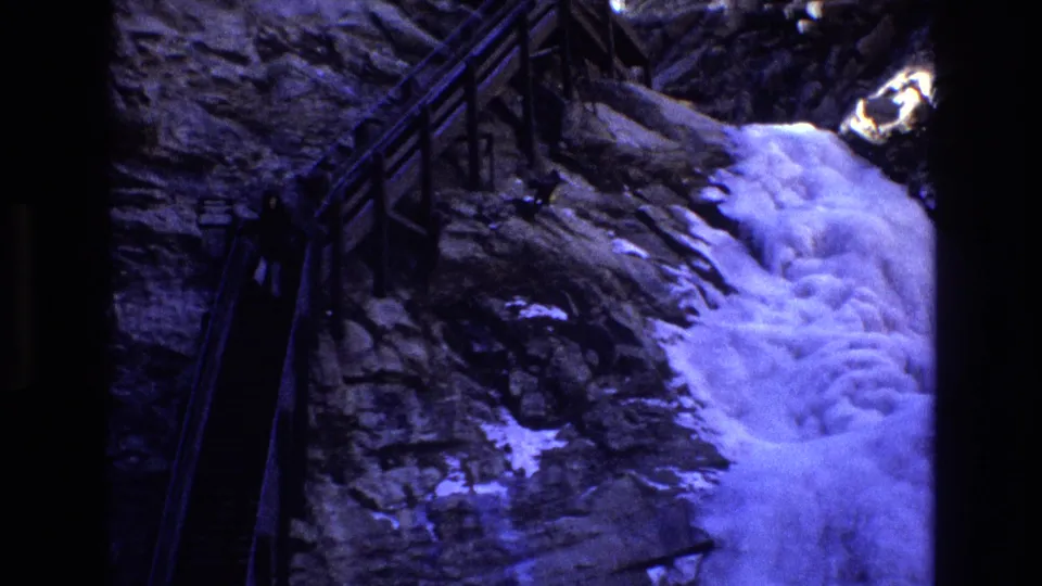 1973 Colorado 8mm frame showing a distant person on a shadowed railed mountain stairway near snow patches and a waterfall on the rocky slope.