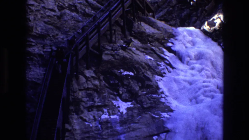 1973 Colorado frame with foaming water dropping along a rock face, snow clinging nearby, and a dark wooden trail railing at the edge of the scene.