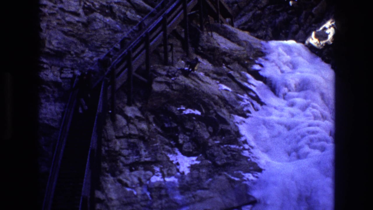 1973 Colorado 8mm frame showing a steep metal-railed stairway along a rugged rock face with a pale waterfall or snow-bright chute nearby.
