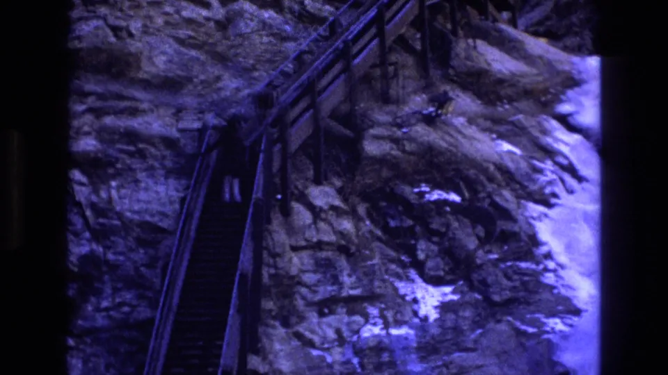 1973 Colorado 8mm frame with the staircase shifted to the left of the rocky cliff, showing railings, rough stone, and a bright chute at the right edge.