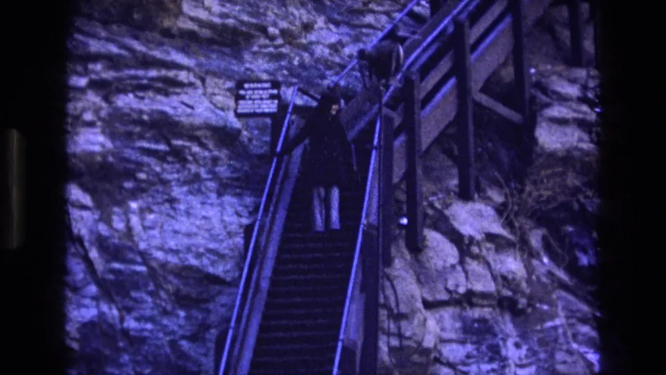 1973 Colorado 8mm frame showing a tourist ascending the cliffside staircase near a small posted sign, with railings and layered rock surrounding the path.