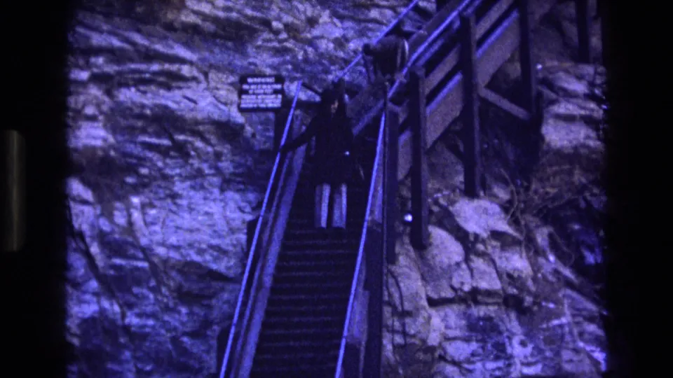 In a 1973 Colorado home-movie frame, a woman in a dark coat and light pants is seen on steep metal stairs beside a rock wall with a small posted sign.