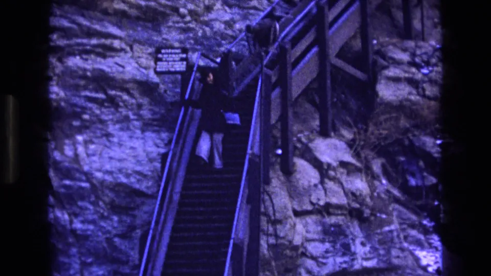 The woman continues down the steep stair flight in this 1973 Colorado 8mm frame, with dark railings, rock surfaces, and a small sign visible nearby.