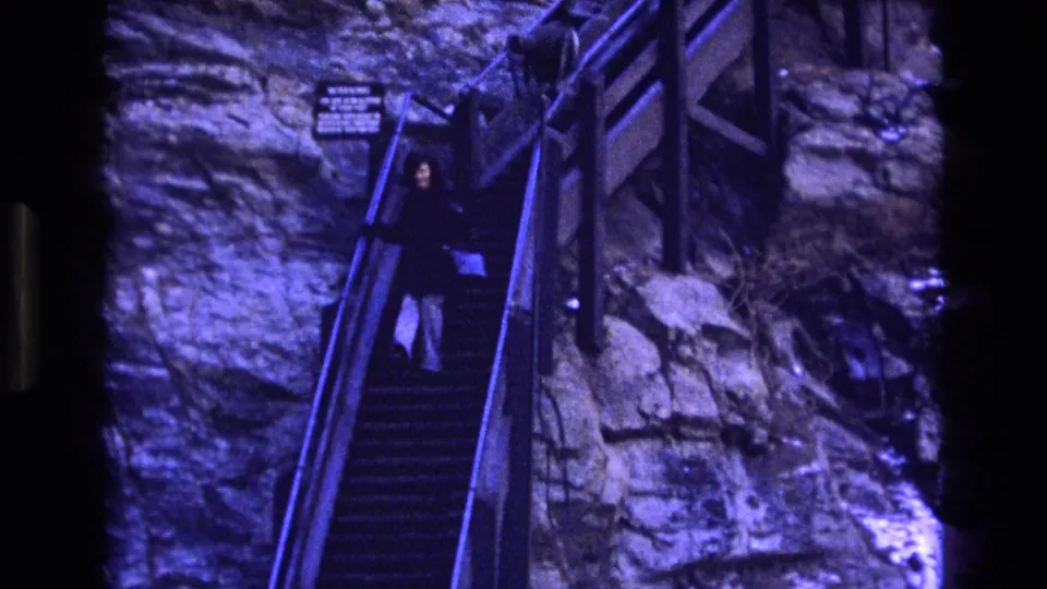 A woman carrying a purse moves farther down the steep stairs, framed by the rocky cliff and railings in blue-purple aged 8mm color.