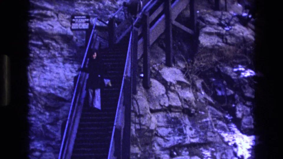 1973 Colorado 8mm frame showing an adult descending a steep outdoor staircase with handrails along a rugged rock face.