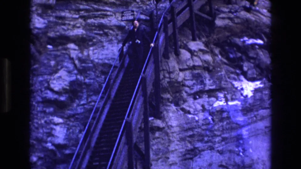 Home-movie frame showing an adult partway down a steep railed stairway beside exposed rock, with a small sign near the upper landing.
