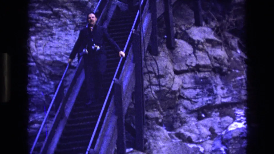 The visitor holds the handrail while moving down the narrow staircase along a rugged rock face in Colorado archival home-movie footage.