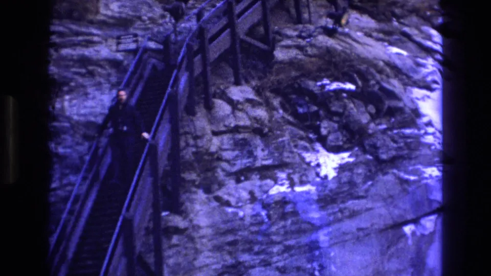 The man appears farther down the rocky outdoor staircase, framed by metal railings and exposed stone in the 1973 Colorado home movie.