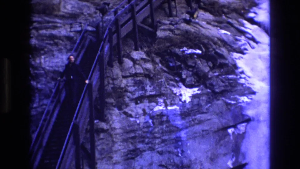 1973 Colorado 8mm frame showing an adult in dark winter clothing descending a steep stairway built along a rocky, snowy cliffside.