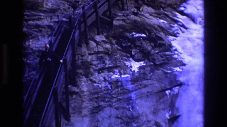 1973 Colorado home-movie frame with a bundled adult moving down a narrow handrailed stairway against a rugged rock face with snow nearby.