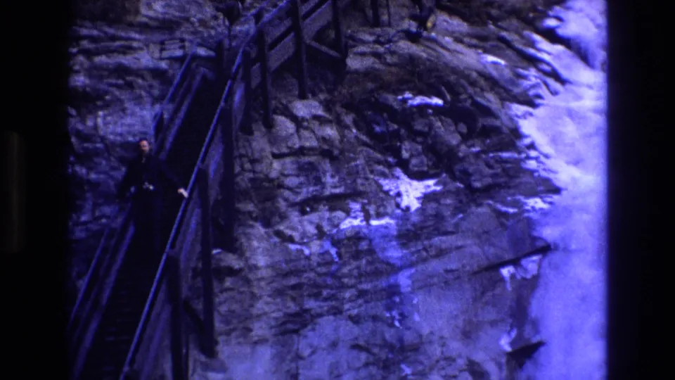 1973 Colorado 8mm frame showing a distant adult on steep stairs with railings, layered rock, snow patches, and strong blue-purple film color shift.