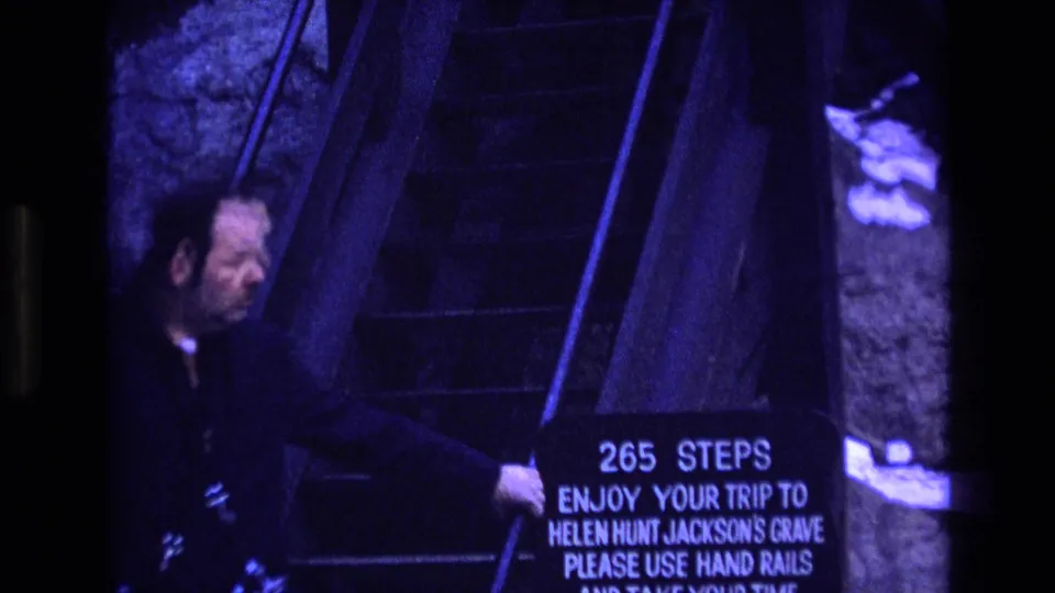 1973 Colorado home-movie frame showing an adult near the base of a dark stairway and a sign that reads 265 steps with instructions to enjoy the trip and use hand rails.