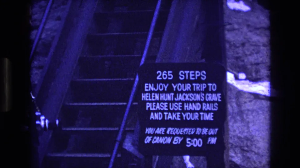 A 1973 Colorado home-movie frame shows the stairway sign for Helen Hunt Jackson's Grave, with part of the adult visitor visible along the left edge.