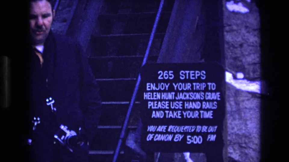 An adult man holding a camera stands near the posted 265 steps sign and the stone stairway to Helen Hunt Jackson's Grave in 1973 Colorado footage.