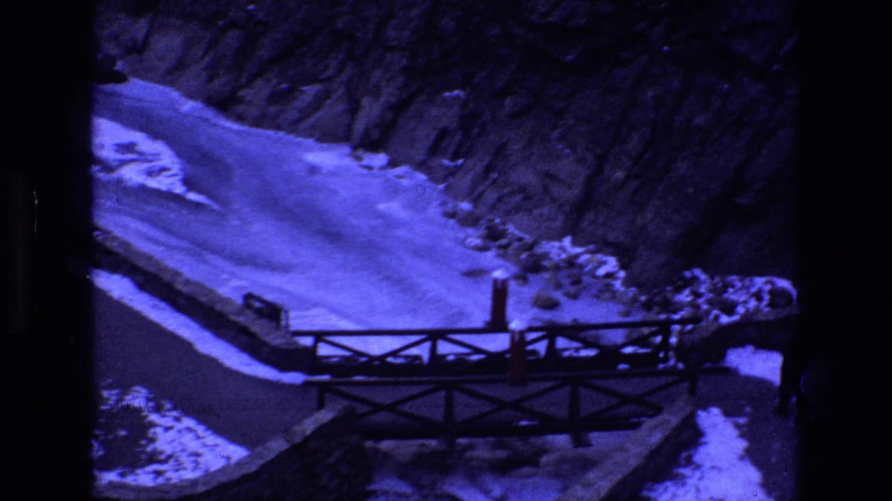 1973 Colorado 8mm frame showing a man in dark winter clothing near a wooden bridge crossing a snow-covered ravine with rocky slopes behind him.
