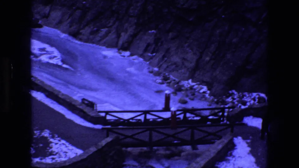 1973 Colorado 8mm frame showing a man in dark winter clothing near a wooden bridge crossing a snow-covered ravine with rocky slopes behind him.