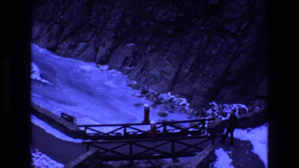 1973 Colorado home-movie frame with a distant man walking at the right end of a wooden bridge over snow and ice below dark rock walls.