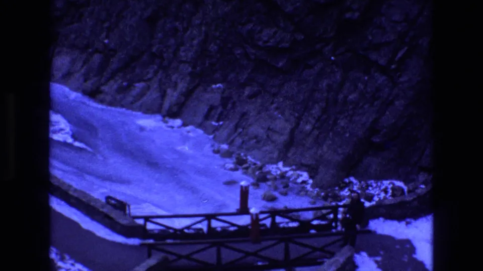 Blue-tinted 1973 Colorado 8mm frame showing the wooden bridge, snowy ravine floor, rocky hillside, and a small dark figure near the right side of the span.