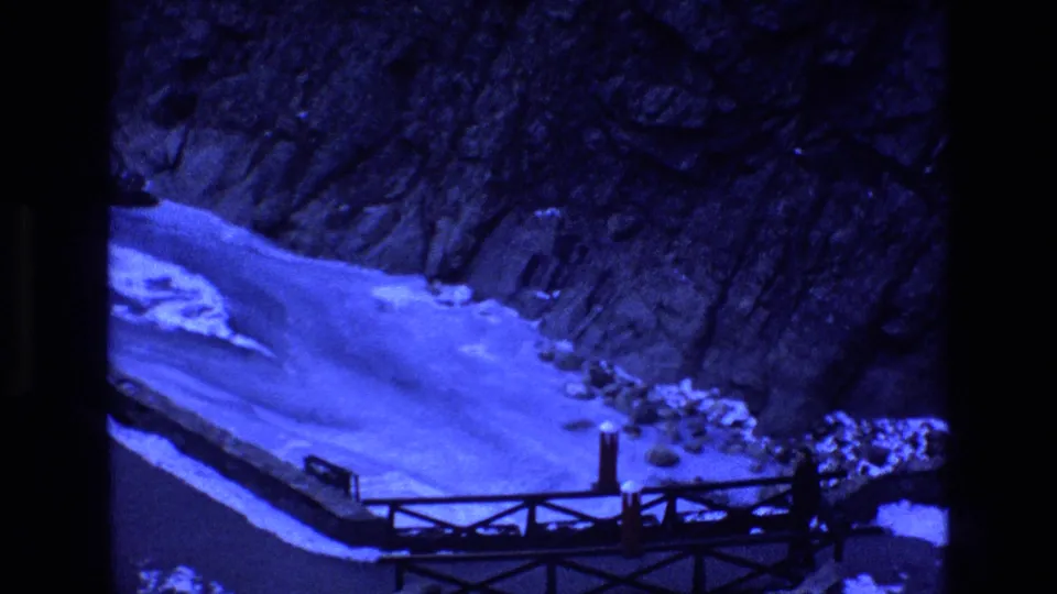 1973 Colorado frame showing a distant man near the right end of a rustic wooden bridge, with snow, ice, and steep rock terrain filling the background.