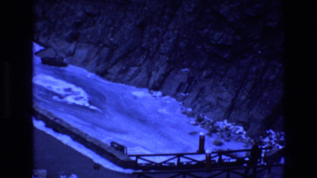 1973 Colorado 8mm frame showing a blue-tinted snowy river canyon, rocky slopes, a wooden overlook railing, and small distant visitor figures.