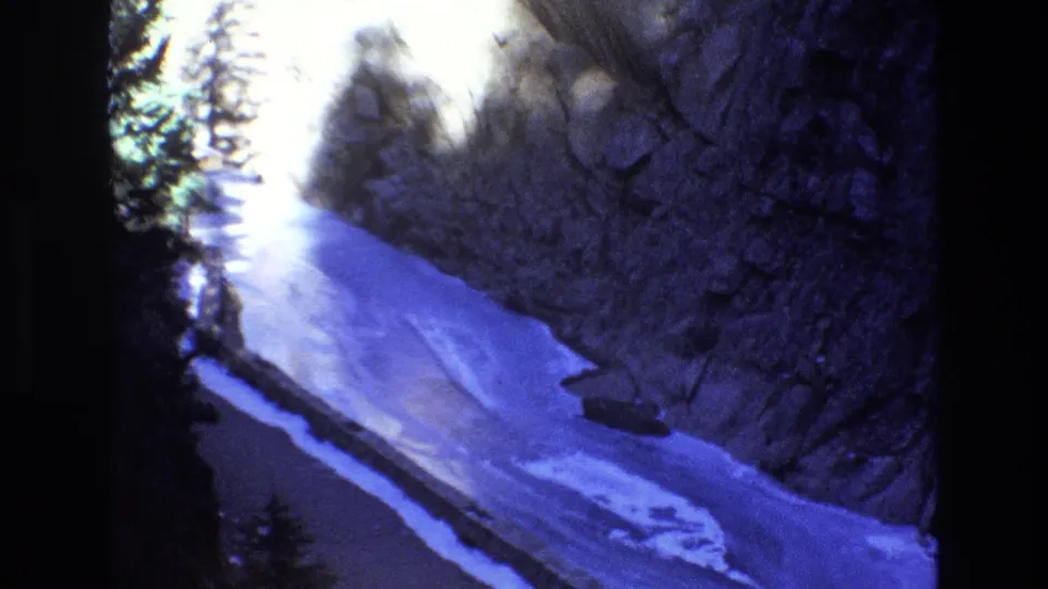 1973 Colorado 8mm frame showing the panned view down a snowy river canyon, with a bright opening at left, rocky wall, trees, and blue-tinted film color.
