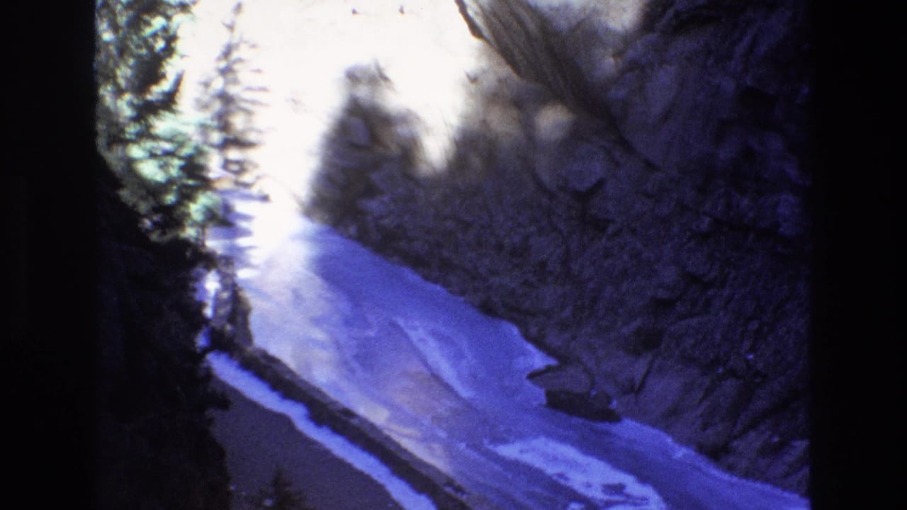 1973 Colorado 8mm frame showing a narrow stream or river running through a shadowed rocky canyon with pine trees along the slope.