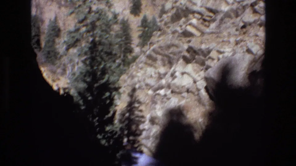 1973 Colorado 8mm frame showing sunlit canyon rocks, scattered pine trees, dark foreground shadow, and a small section of stream near the lower edge.