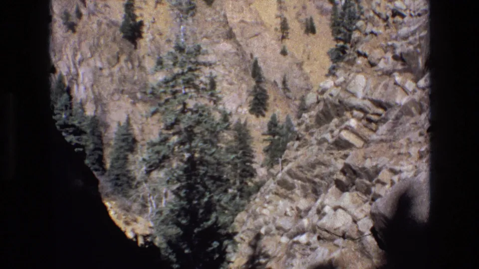 1973 Colorado 8mm frame showing a rocky canyon wall with scattered pine trees, strong sunlight, dark vignetted edges, and little visible sky.