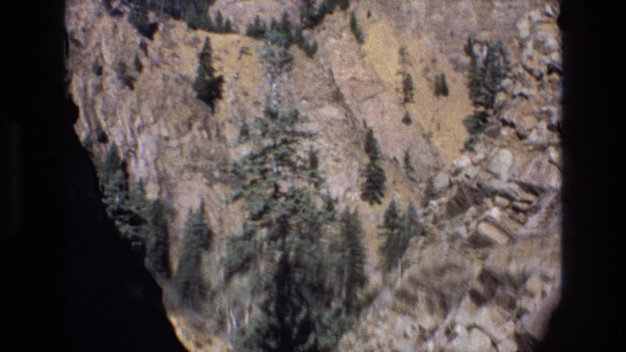 1973 Colorado 8mm frame showing a rocky mountain slope with scattered evergreen trees, dry scrub, blue sky, and dark gate edges from the home-movie scan.